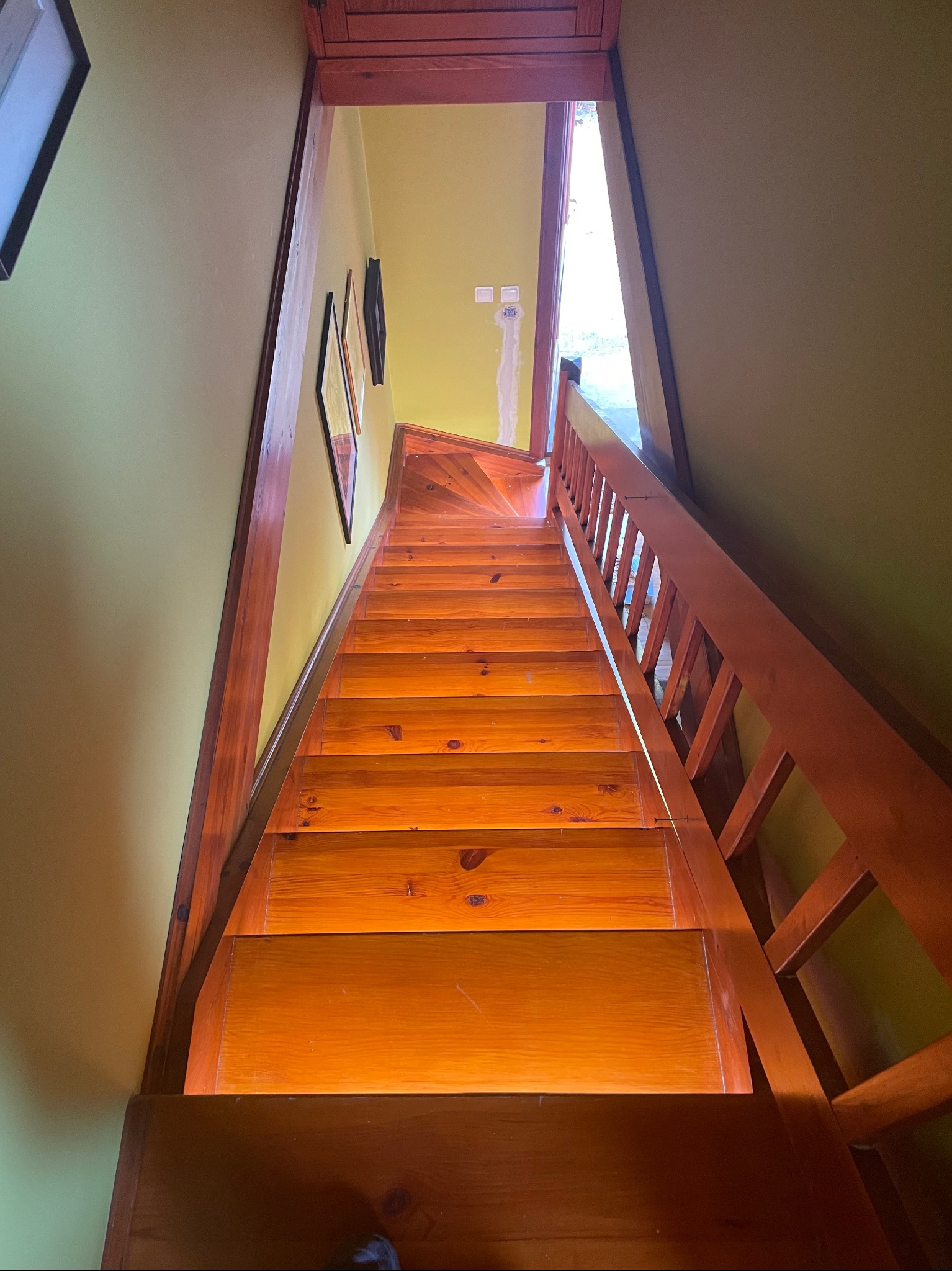 Wysokie ujęcie drewnianych schodów z balustradą w domu o ścianach w odcieniach zieleni i żółci, widoczne gniazdka elektryczne na ścianie przy górnym biegu schodów.