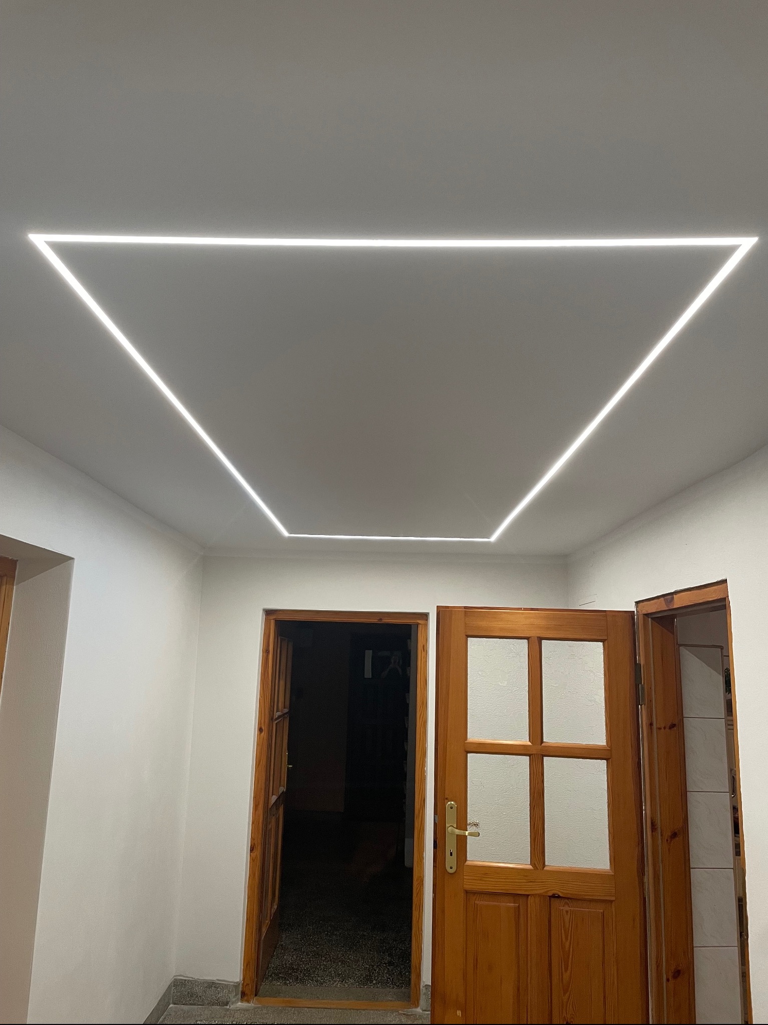 Nowoczesne oświetlenie sufitowe LED w kształcie trójkąta, z widocznymi drewnianymi drzwiami z szybami w tle.