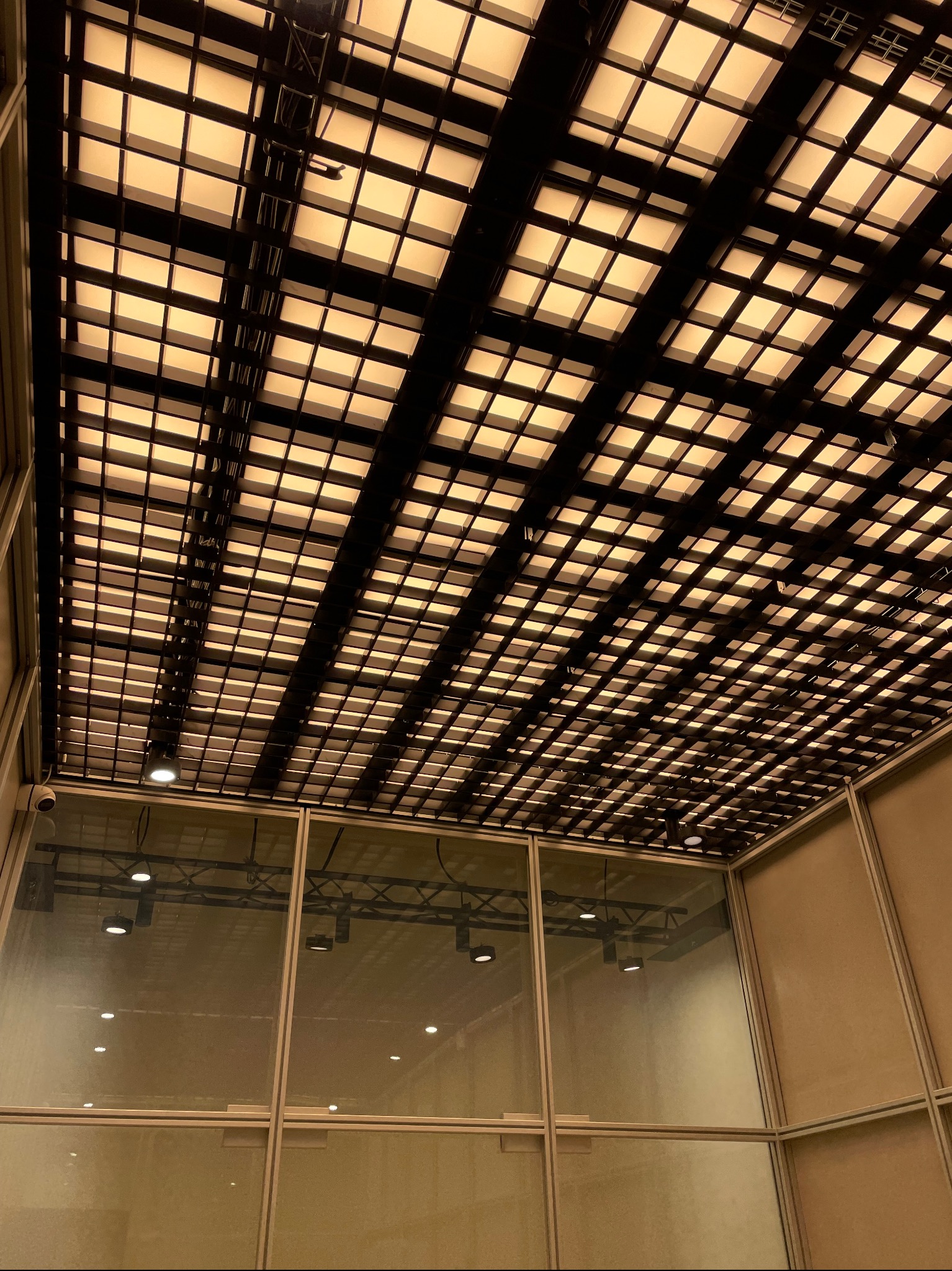 Sufit z rastrem oświetleniowym w pomieszczeniu biurowym, widoczne lampy LED w kwadratowych panelach, kamera monitoringu w rogu.