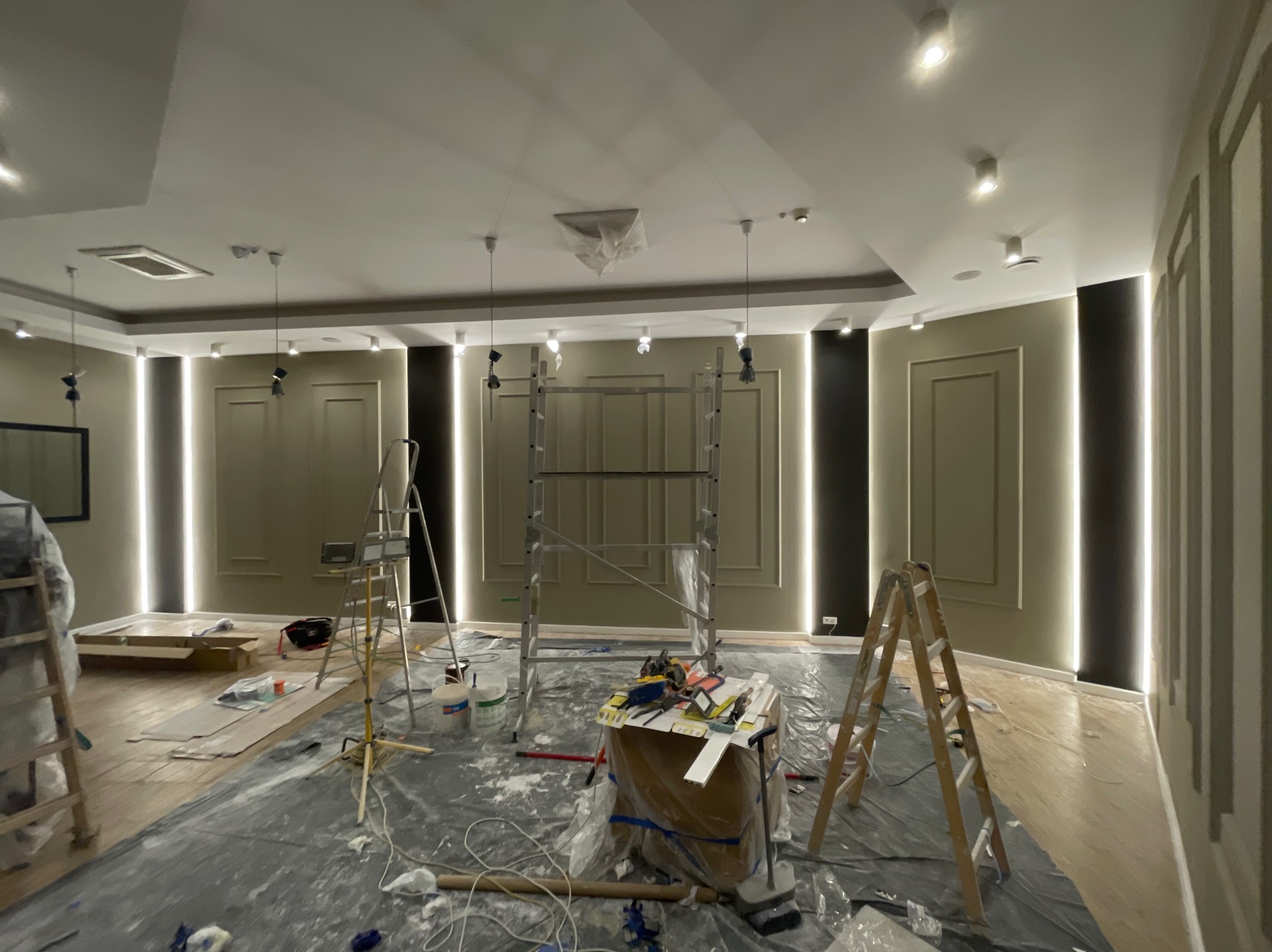 Wnętrze w trakcie remontu z zamontowanym oświetleniem LED wzdłuż ścian, widoczne rusztowania, drabiny, narzędzia i zabezpieczona folią podłoga.