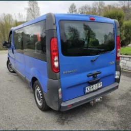 Niebieski bus Renault Trafic z hakiem holowniczym, zaparkowany na asfalcie na tle zieleni i kamiennego murku.