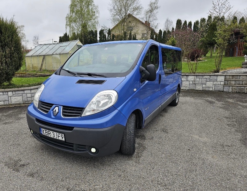 Niebieski bus Renault Trafic zaparkowany na podjeździe z kostki brukowej, z widocznym numerem rejestracyjnym KBR 3JF9, na tle zadbanego ogrodu z szklarnią.