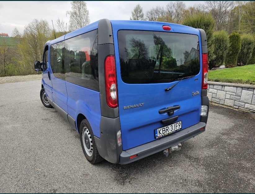 Niebieski bus Renault Trafic z hakiem holowniczym, zaparkowany na asfalcie na tle zieleni i kamiennego murku.