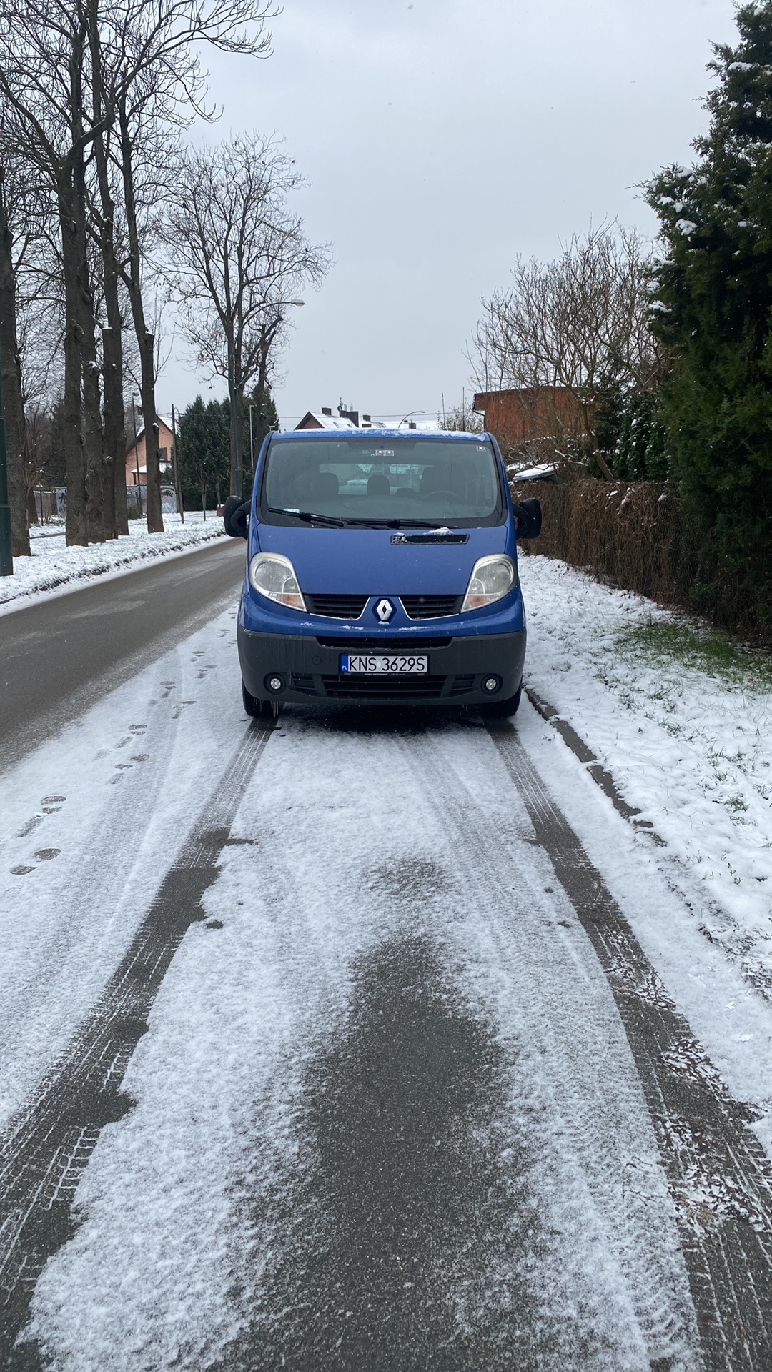 Niebieski bus Renault Trafic zaparkowany na ośnieżonej drodze w pochmurny dzień, widoczne ślady opon na śniegu.
