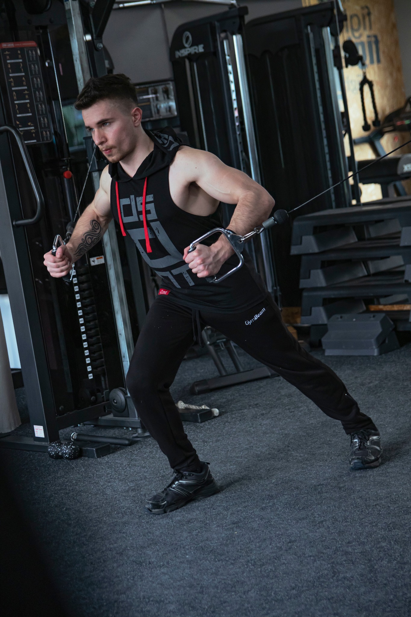 Młody mężczyzna w czarnym stroju sportowym ćwiczy na maszynie z linkami w siłowni, widoczny tatuaż na ramieniu.