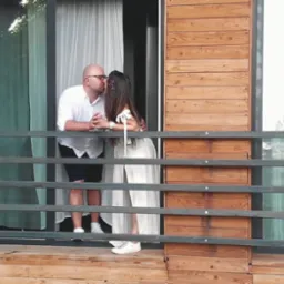 Para młoda w białych strojach całuje się na drewnianym balkonie z metalową balustradą, widok z dołu.