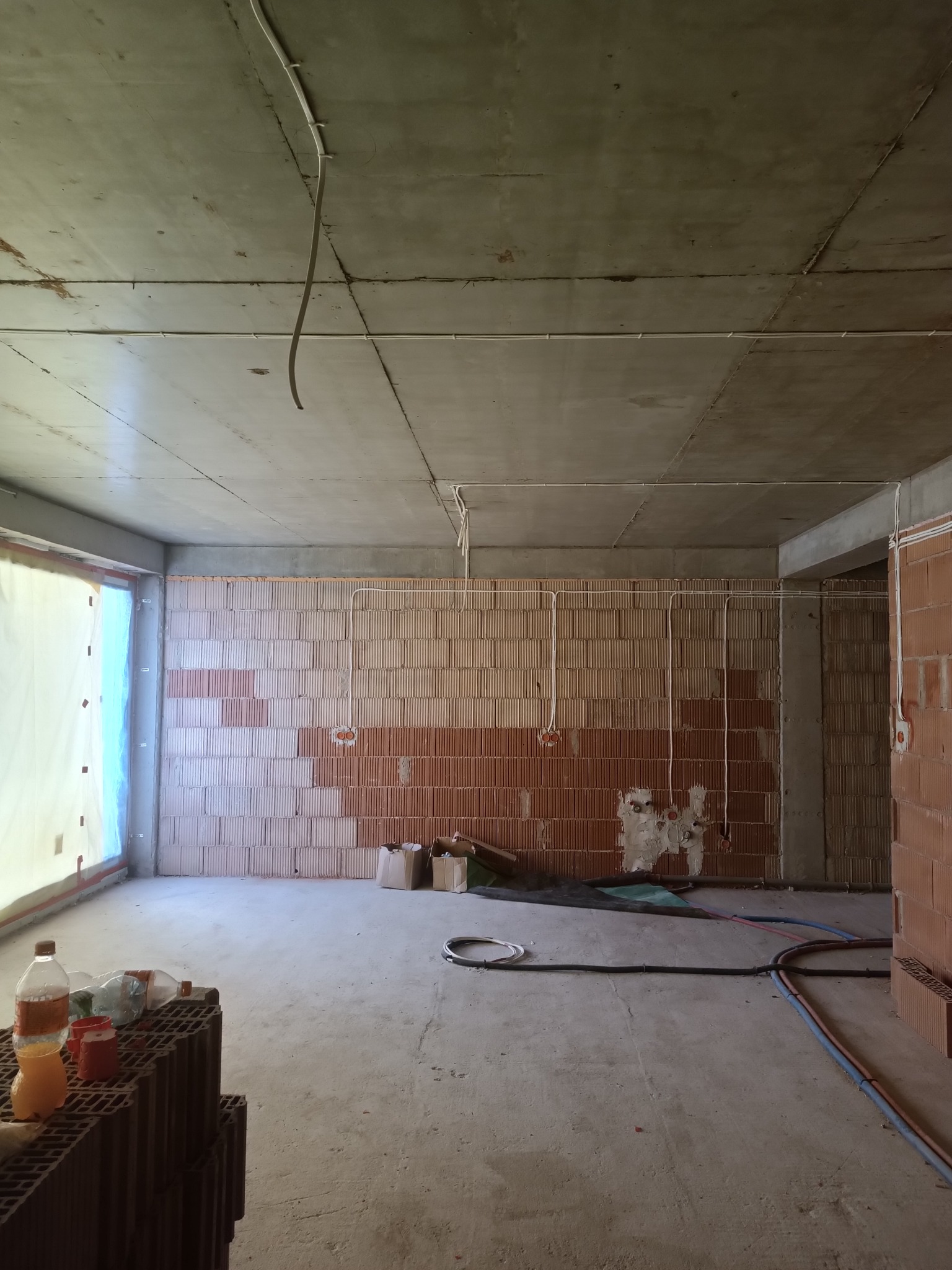 Surowy stan budynku w trakcie prac instalacyjnych: widoczne przewody elektryczne prowadzone po ścianach z pustaków ceramicznych, okno zabezpieczone folią, podłoże betonowe, butelki i kartony...