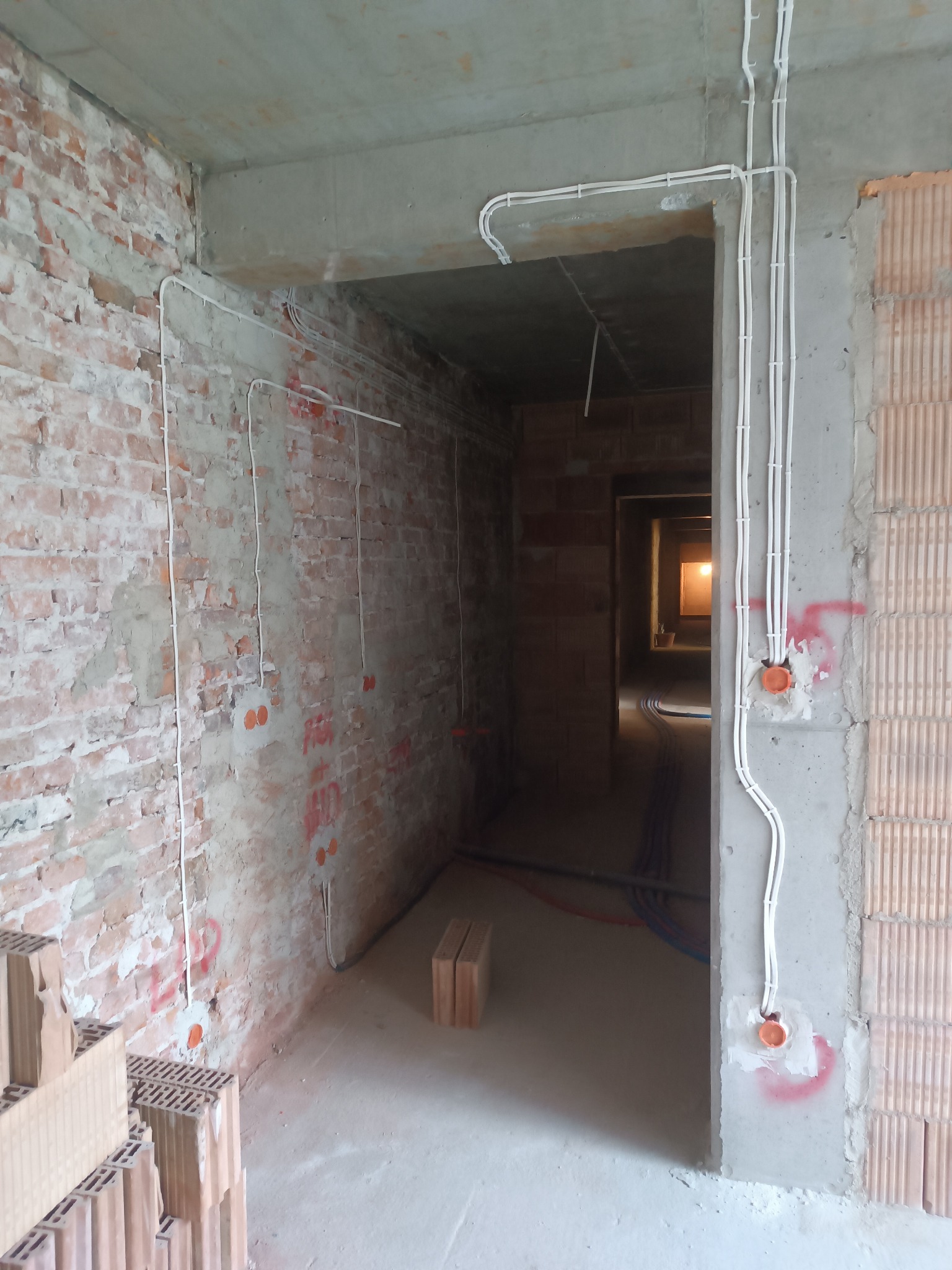Surowy stan instalacji elektrycznej w budynku w trakcie budowy: widoczne białe przewody poprowadzone po ceglanej ścianie i betonowych elementach konstrukcyjnych, pomarańczowe puszki elektryczne...