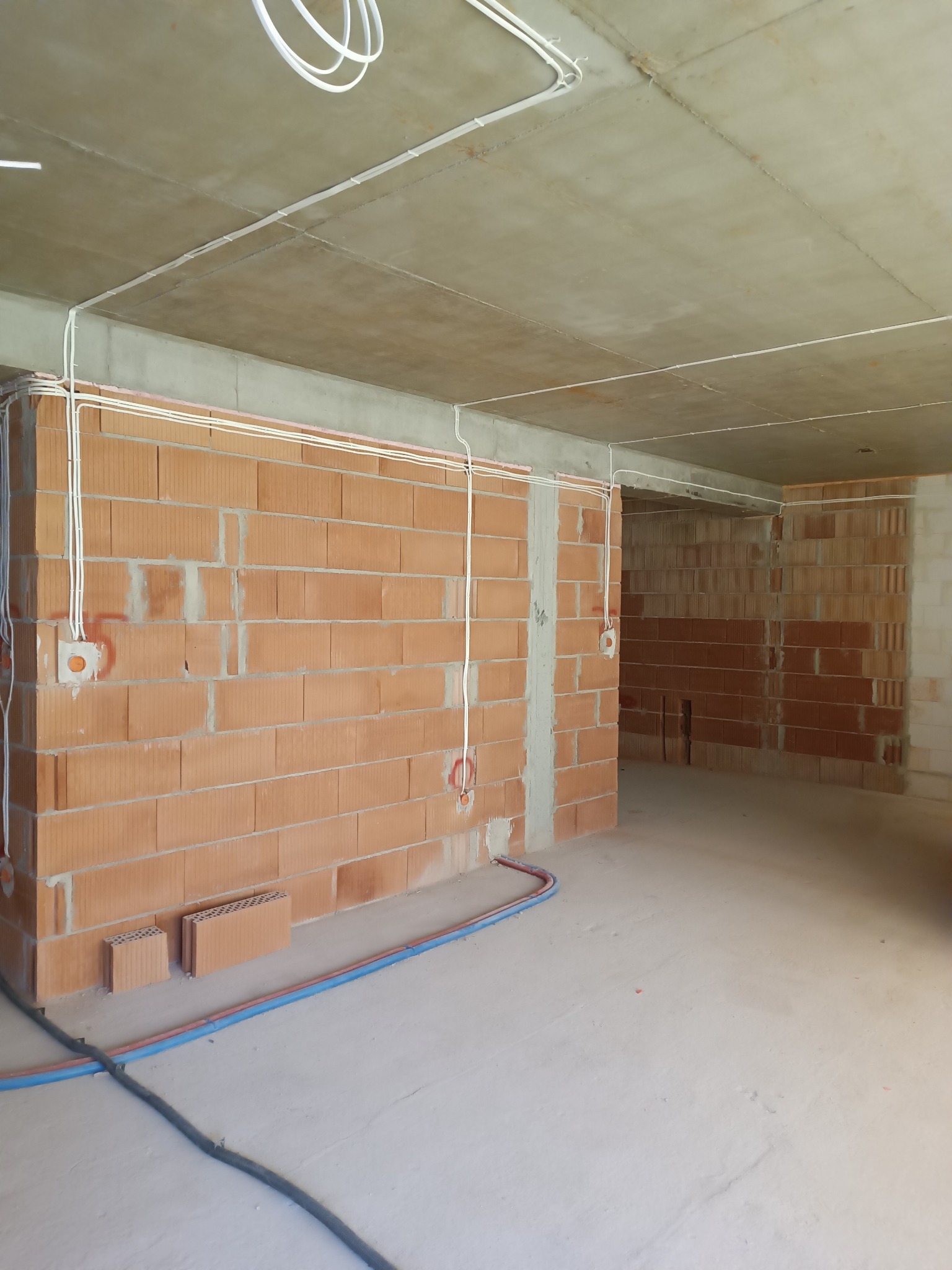 Wnętrze w stanie surowym w Błesznie z widoczną instalacją elektryczną: kable prowadzone po ścianach z cegły i pod sufitem, puszki elektryczne w ścianach.