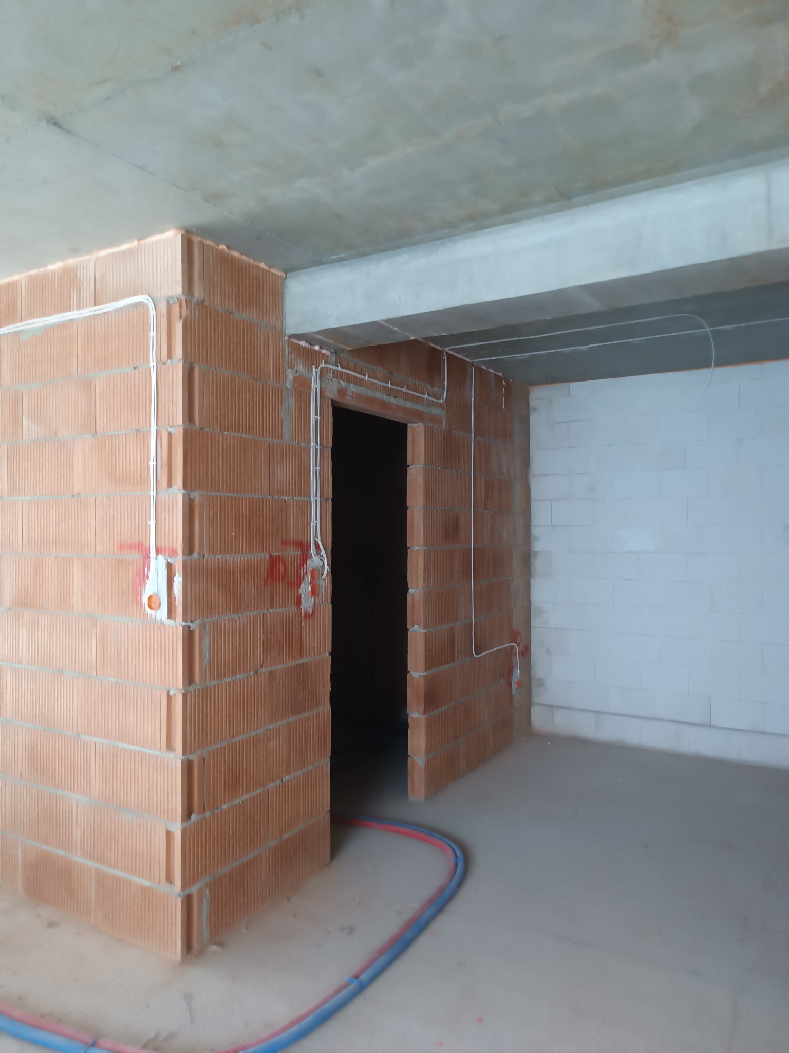 Surowy stan: Instalacja elektryczna w nowym budynku z czerwonej cegły, widoczne białe kable w peszlach, otwory pod puszki elektryczne zaznaczone czerwoną farbą, szare ściany i betonowy sufit.