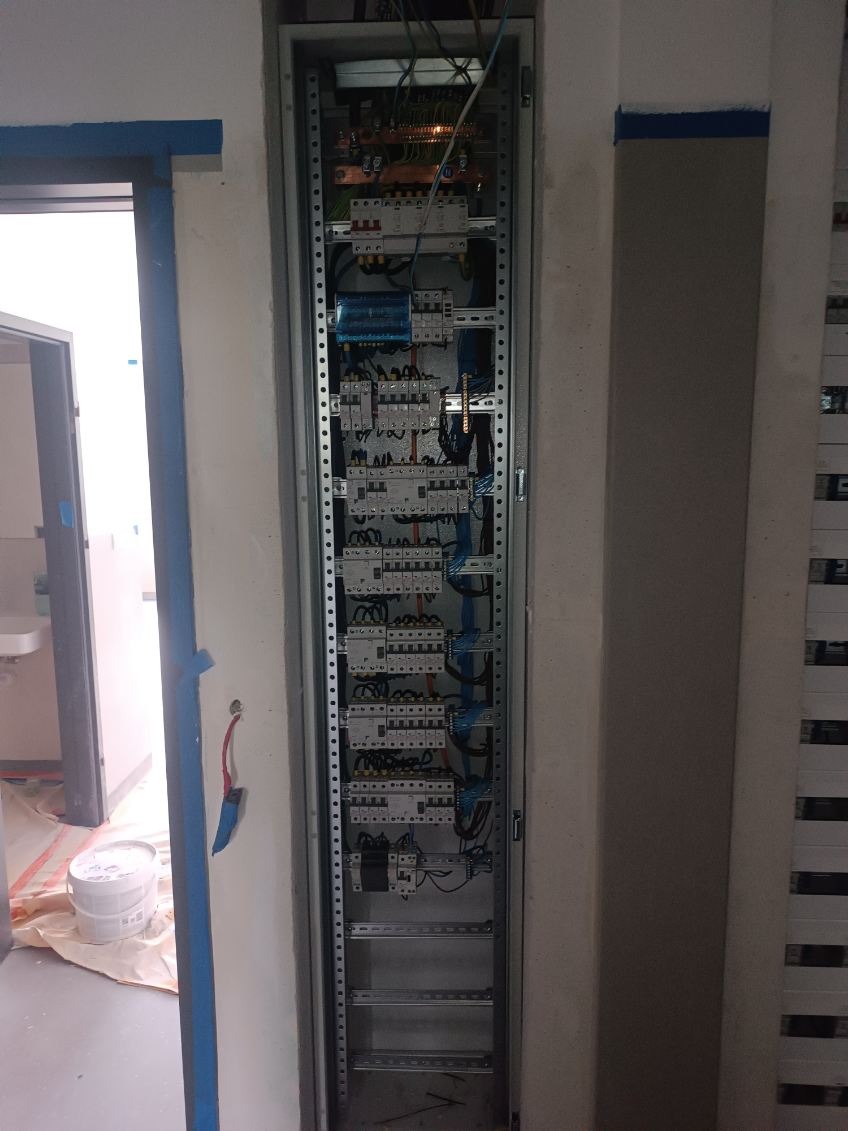 Rozdzielnica elektryczna z zabezpieczeniami zamontowana w wąskiej wnęce w trakcie prac wykończeniowych, widoczne przewody i aparatura zabezpieczająca.