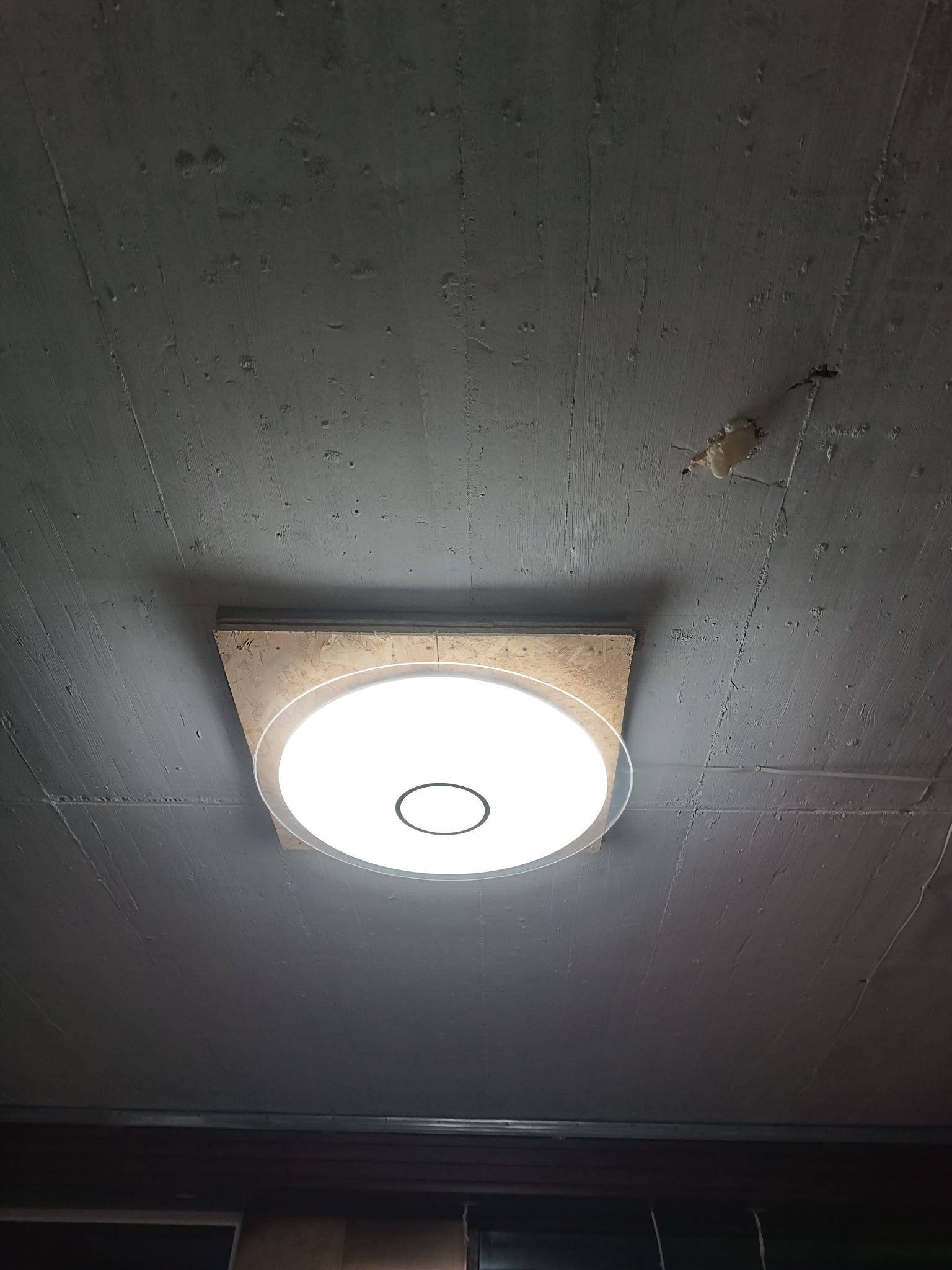 Zamontowana okrągła lampa sufitowa LED, przymocowana do płyty OSB na nierównym, szarym suficie z widoczną pianką montażową.