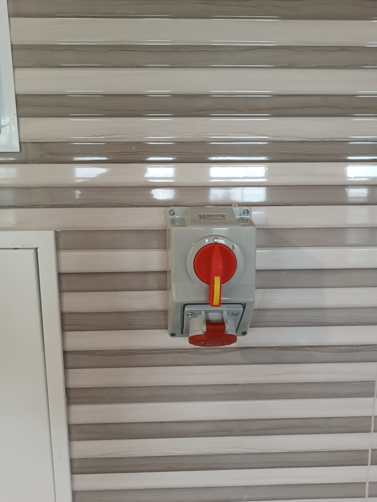 Szary wyłącznik prądu z czerwoną dźwignią i gniazdem, zamontowany na ścianie z poziomymi panelami w odcieniach beżu i szarości.