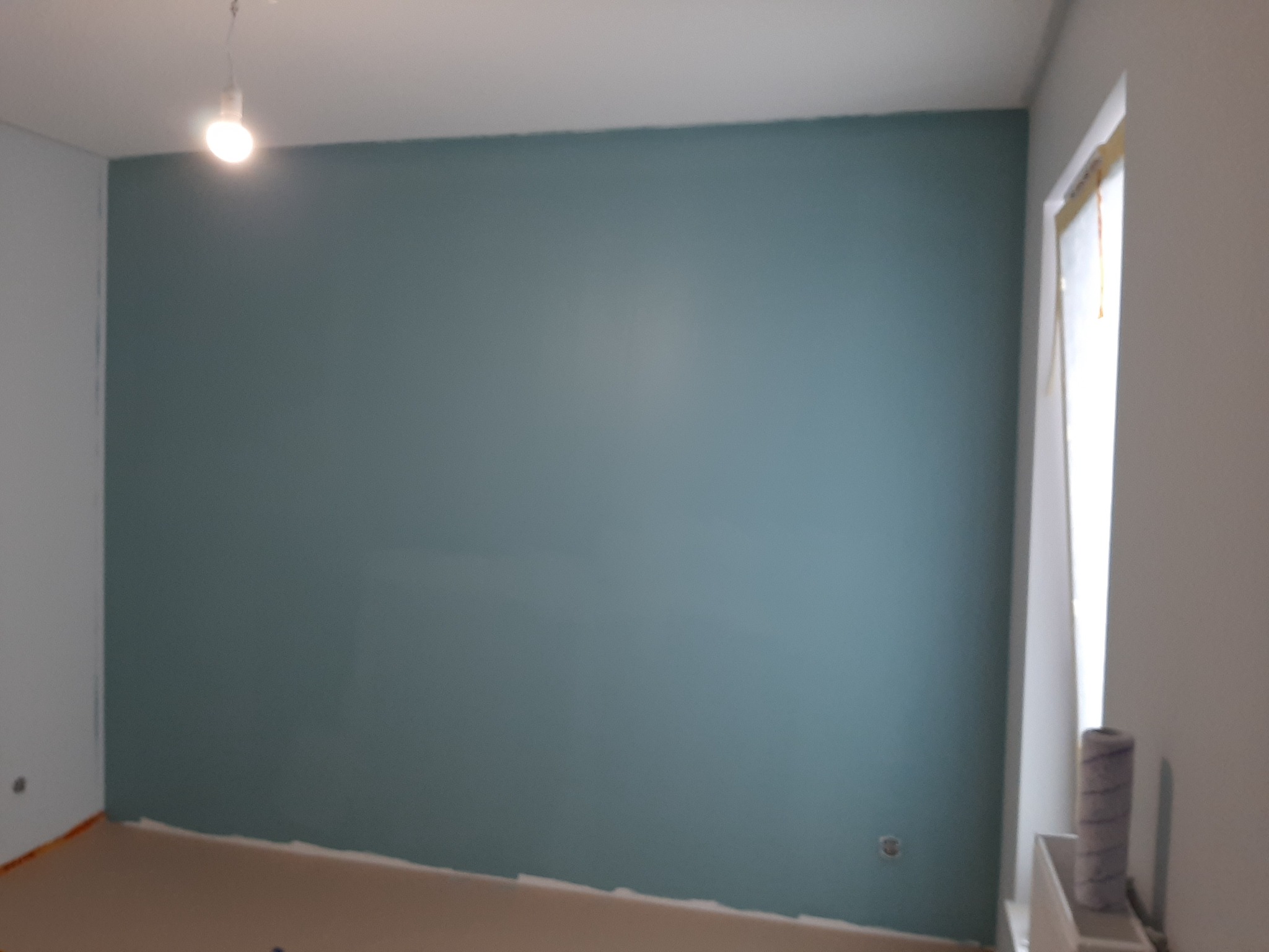 Pomieszczenie po malowaniu na kolor turkusowy, widoczny wałek malarski, zabezpieczona folią ościeżnica okienna i podłoga.