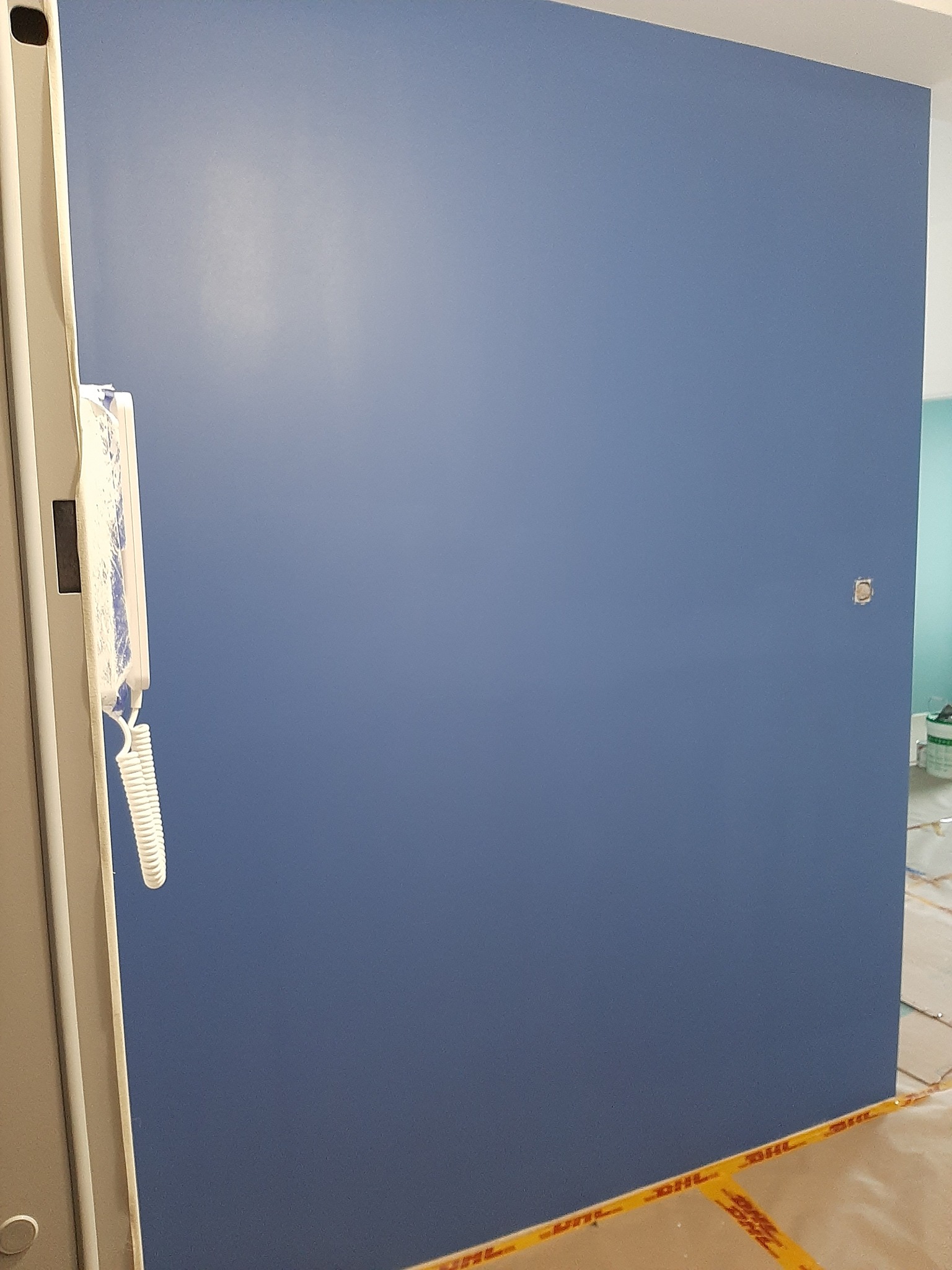 Świeżo pomalowana na niebiesko ściana z widocznym domofonem po lewej stronie, zabezpieczona taśmą malarską na podłodze.