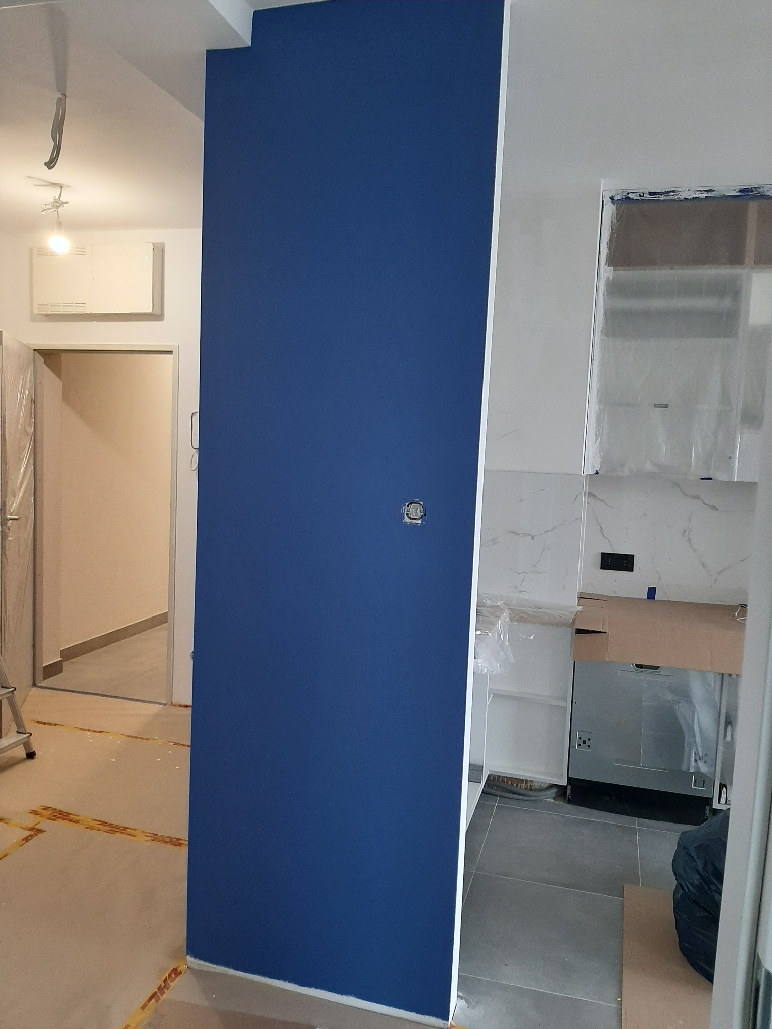 Ściana pomalowana na intensywny niebieski kolor w mieszkaniu w Łodzi, widoczny fragment kuchni z zabezpieczonymi folią meblami i marmurowymi płytkami, podłoga zabezpieczona kartonem.