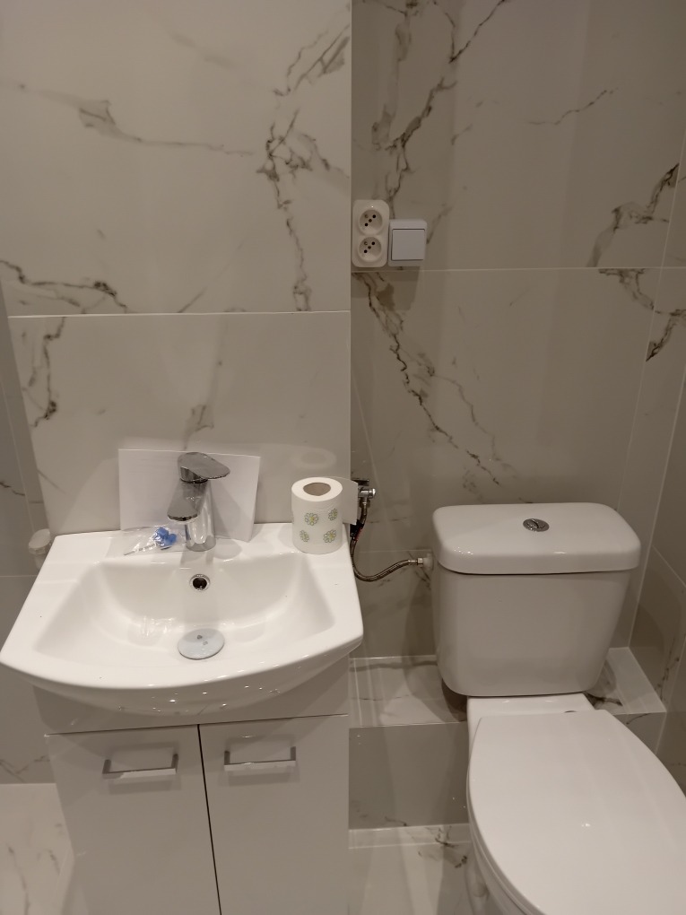 Biała umywalka z szafką, toaleta i marmurowe płytki w wykończonej łazience, widoczne gniazdko elektryczne na ścianie.