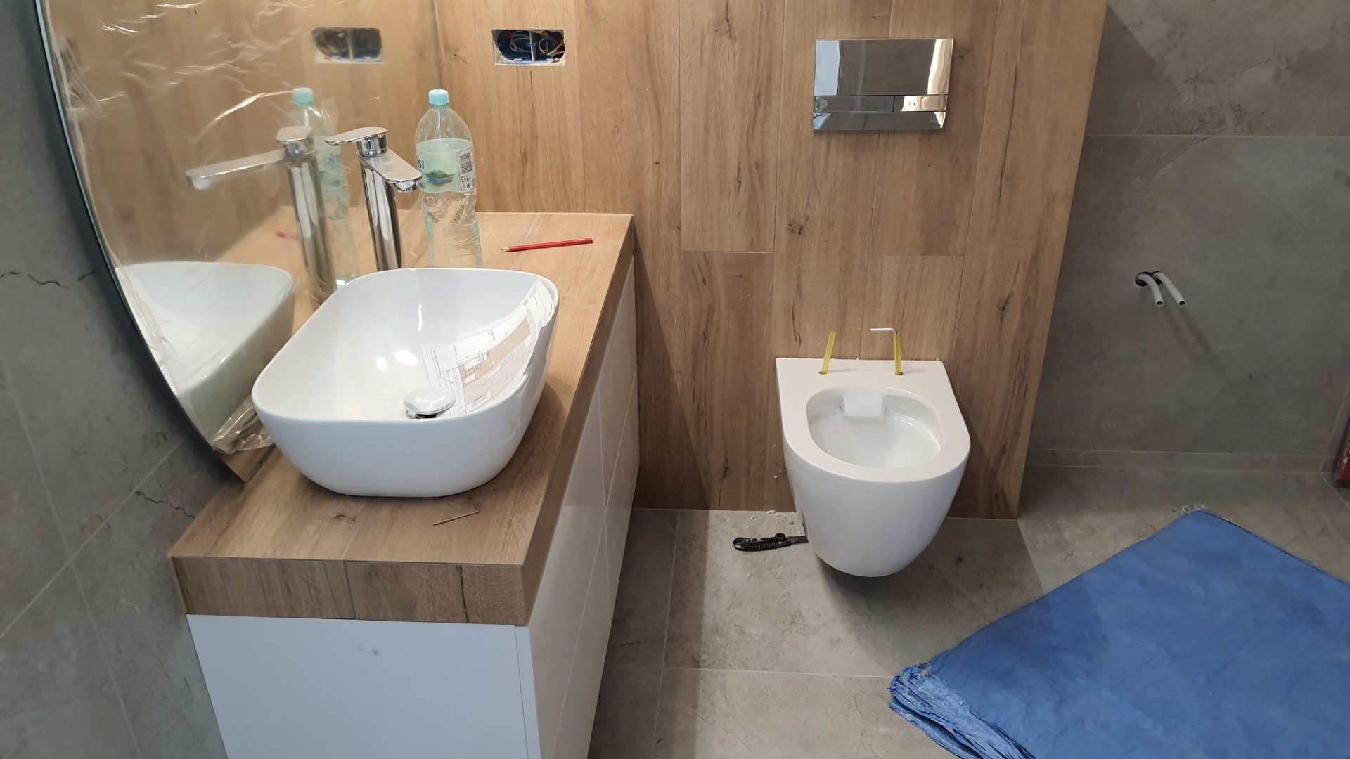 Montaż wiszącej miski WC i umywalki nablatowej w łazience wykończonej płytkami imitującymi drewno i szary kamień, widoczne niezabezpieczone przewody elektryczne.
