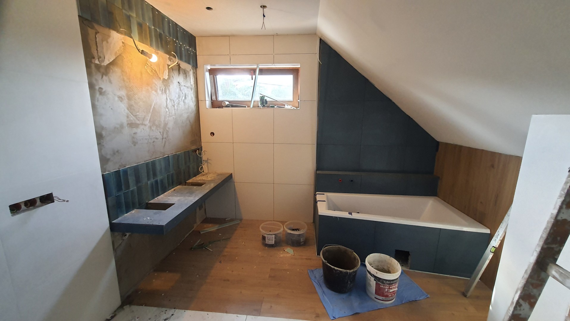 Remontowana łazienka na poddaszu: położone płytki w strefie umywalki i wanny, widoczne niedokończone prace wykończeniowe, wiadro na podłodze.