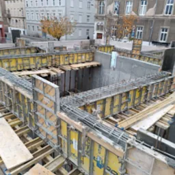 Fundamenty domu w trakcie budowy z widoczną konstrukcją szalunkową i zbrojeniem stalowym, w tle zabudowa miejska.