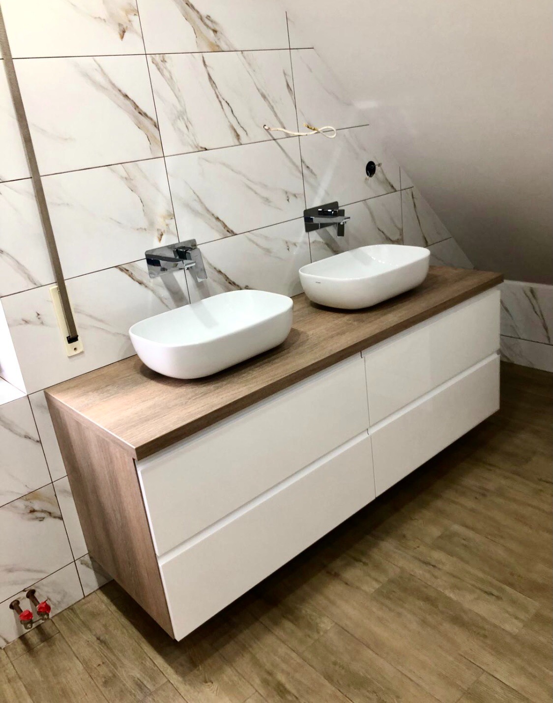 Łazienka na poddaszu: dwie owalne umywalki nablatowe na drewnianym blacie z białymi szafkami, marmurowe płytki na ścianie, chromowane baterie.