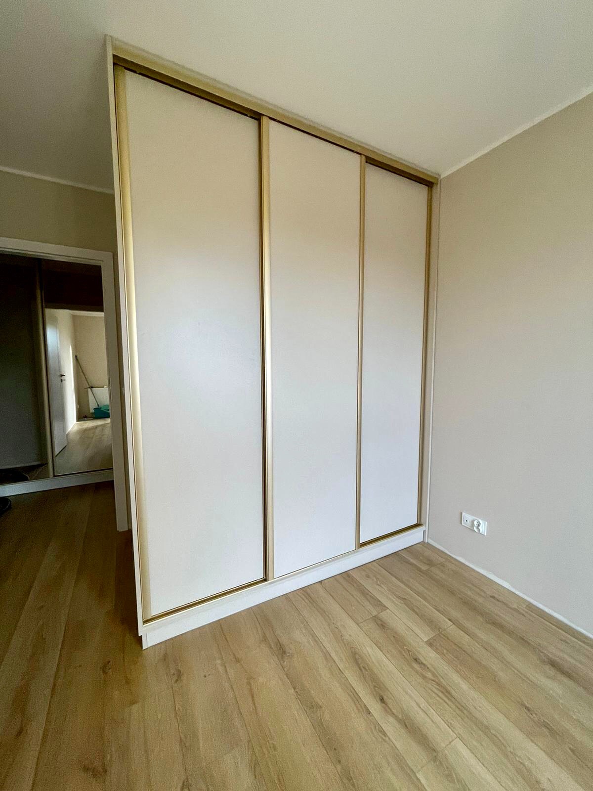 Trzydrzwiowa szafa wnękowa z przesuwnymi drzwiami w kolorze złamanej bieli i złotymi ramami, na podłodze panele w odcieniach beżu, widok na korytarz z lustrem.