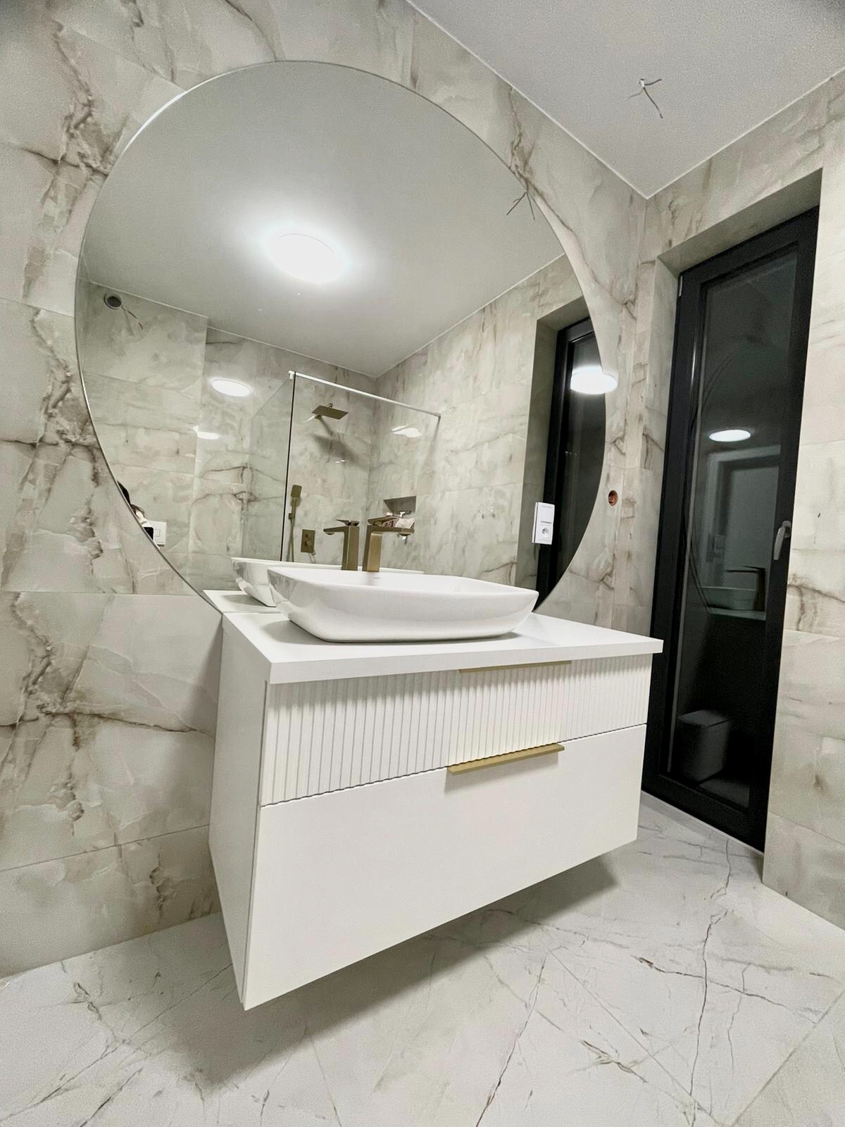 Nowoczesna łazienka z owalnym lustrem odbijającym kabinę prysznicową, biała szafka pod umywalkę z frezowanymi frontami i złotymi uchwytami, marmurowe płytki na ścianach i podłodze.