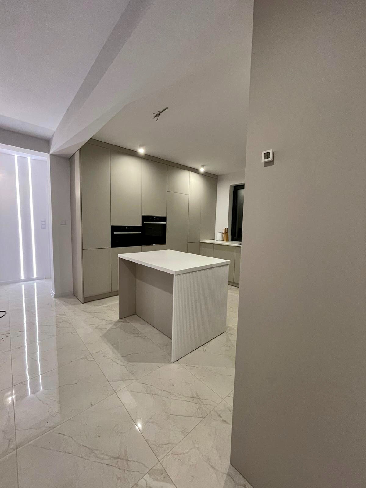 Nowoczesna kuchnia z szarymi szafkami na wymiar i białą wyspą z ryflowanym wykończeniem, marmurowe płytki podłogowe odbijające światło.