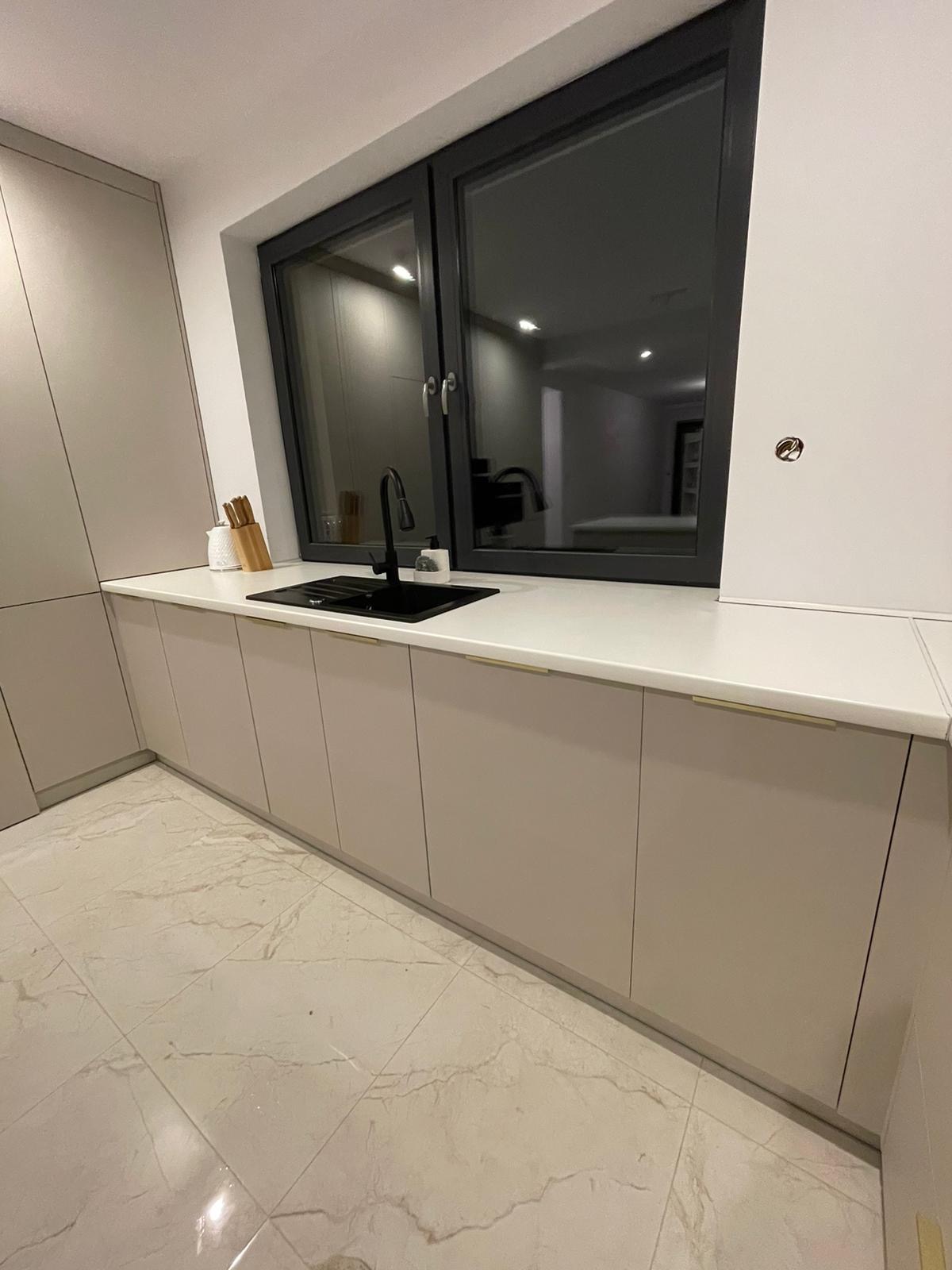 Beżowa zabudowa kuchenna z czarnym zlewem i baterią, biały blat, okno w czarnej ramie, marmurowa podłoga.