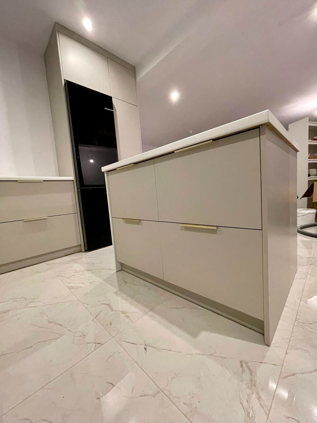 Beżowa wyspa kuchenna z białym blatem i złotymi uchwytami, marmurowa podłoga, zabudowa lodówki w tle.