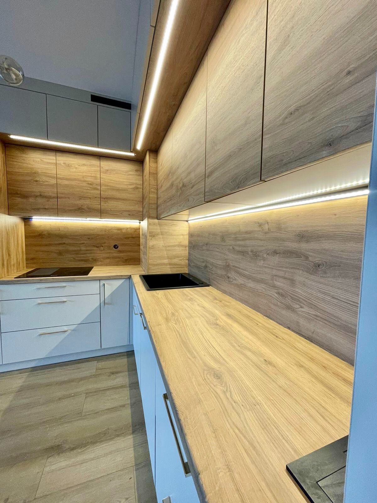 Nowoczesna kuchnia z drewnianymi szafkami górnymi, białymi dolnymi i podświetleniem LED, czarny zlew i płyta indukcyjna, jasna drewniana podłoga.