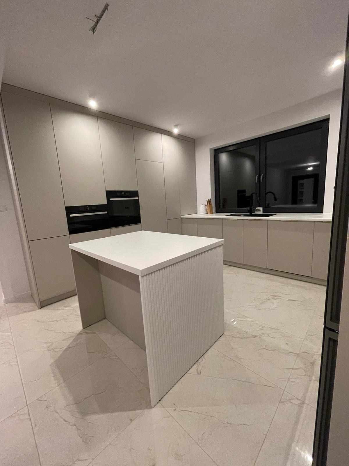 Nowoczesna kuchnia w odcieniach beżu z wyspą z ryflowanym wykończeniem, czarnym oknem i sprzętem AGD w zabudowie.