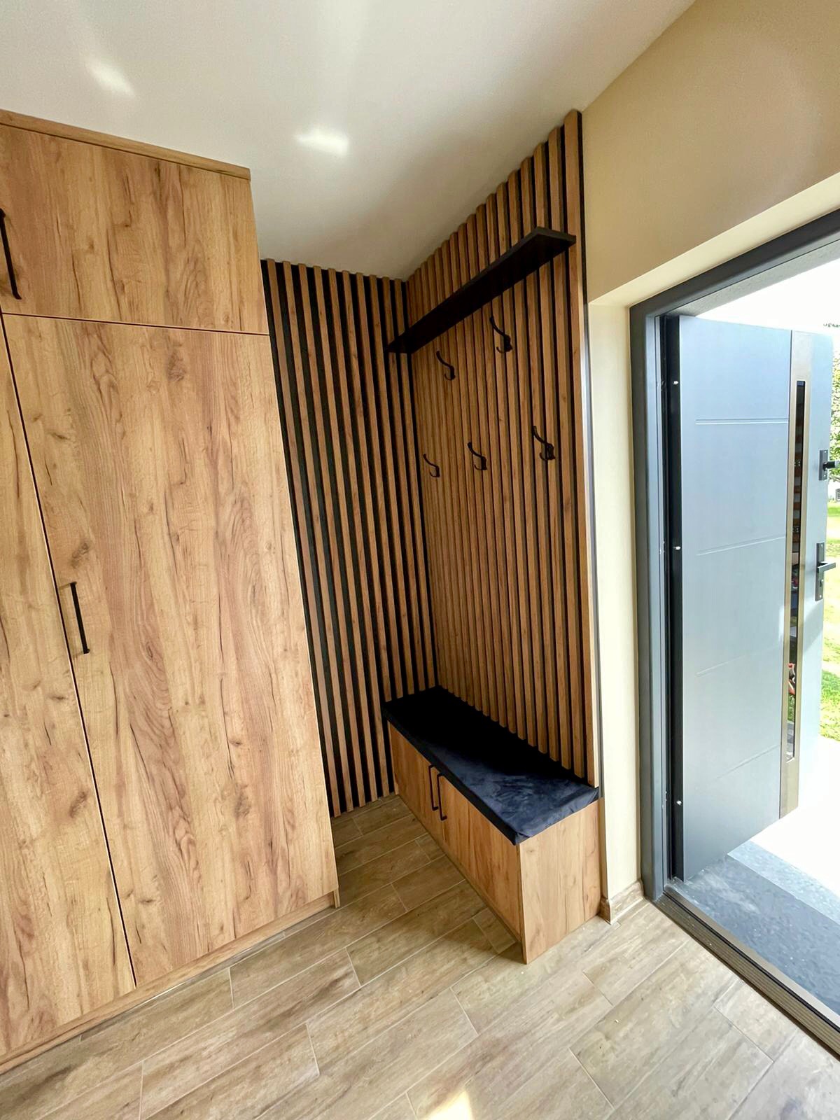Przedpokój z zabudową z drewnianych lameli, szafa z frontami w kolorze drewna, siedzisko z czarną tapicerką i wieszaki na ścianie, nowoczesne drzwi wejściowe.