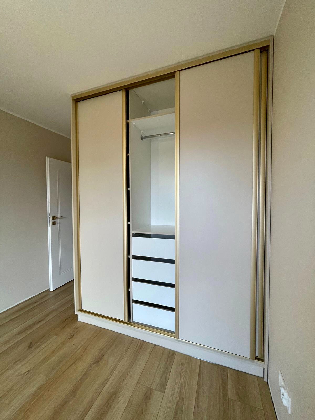 Biała szafa wnękowa z przesuwnymi drzwiami i złotymi obramowaniami, widoczne półki i szuflady, jasne wnętrze, parkiet na podłodze.