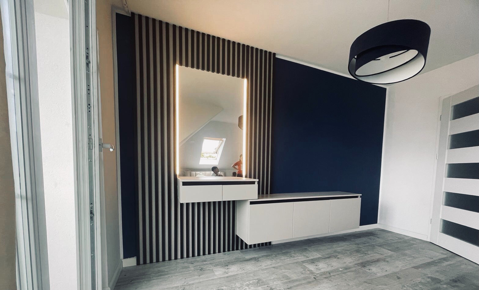Nowoczesny przedpokój z szafkami wiszącymi, lustrem z podświetleniem LED w ramie z drewnianych listew, ściana w kolorze granatowym, szara podłoga imitująca drewno, widoczna lampa sufitowa i białe...