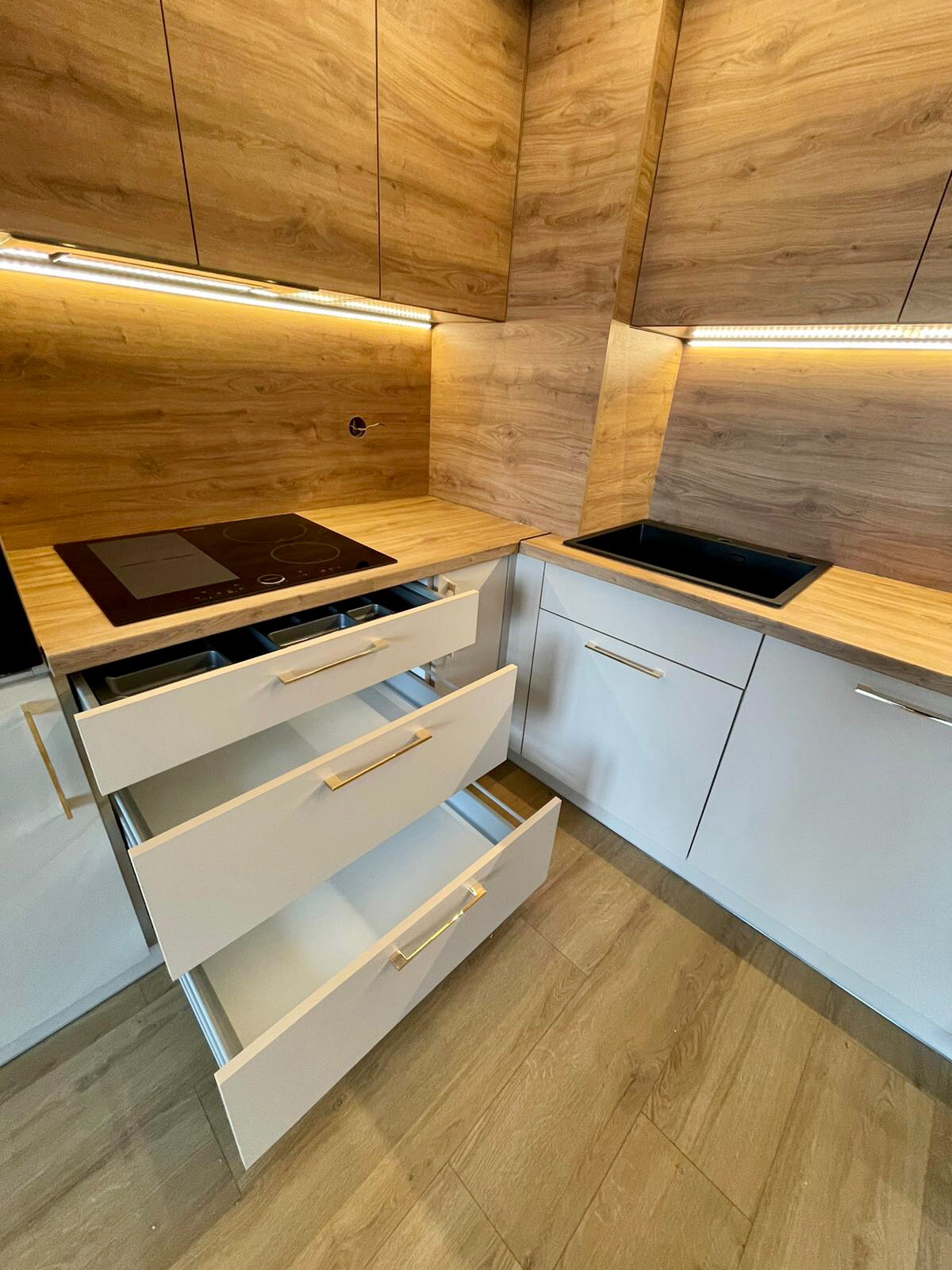 Nowoczesna kuchnia z drewnianymi frontami szafek górnych i blatem, białe szafki dolne z otwartymi szufladami i złotymi uchwytami, czarna płyta indukcyjna i zlew.