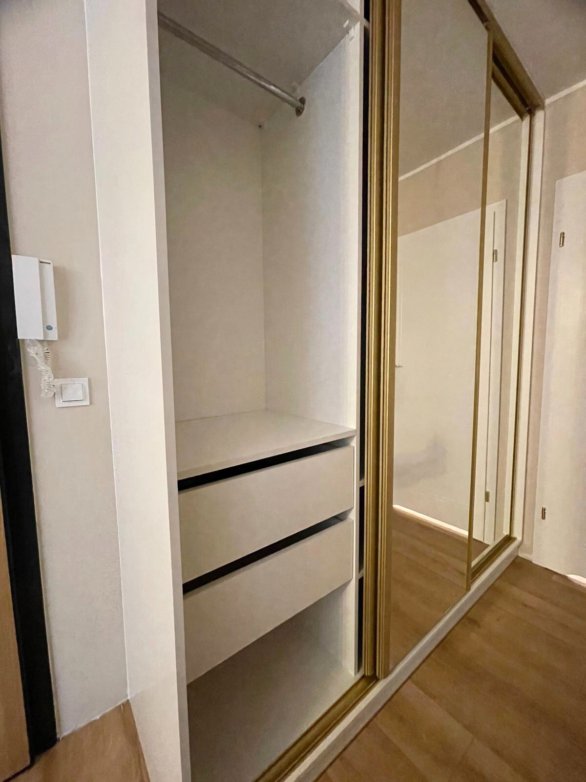 Wnętrze szafy wnękowej z przesuwnymi drzwiami lustrzanymi w złotych ramach, widoczne dwie szuflady i drążek na ubrania, ściana z domofonem.