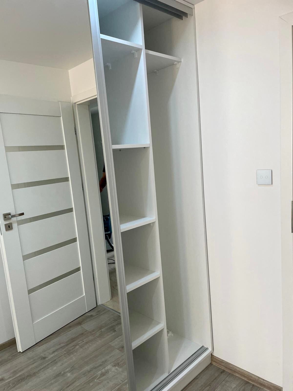 Biała szafa wnękowa z otwartymi drzwiami przesuwnymi, widoczne półki, drzwi pokojowe z poziomymi wstawkami w tle.