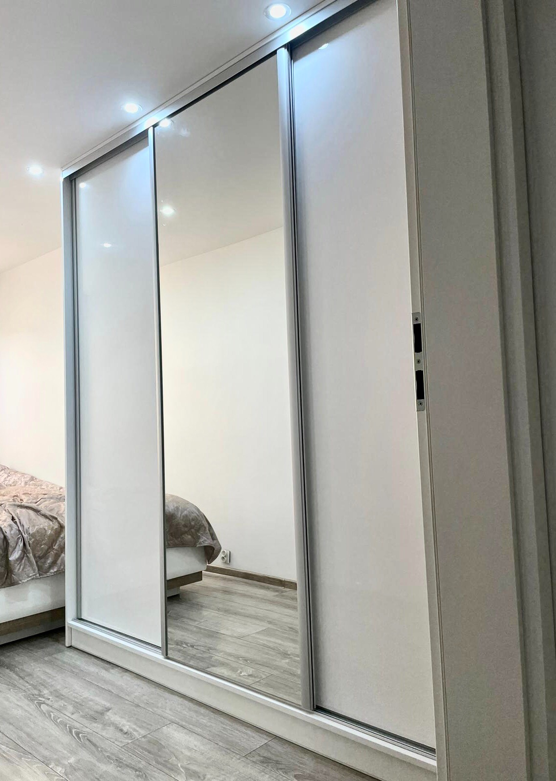 Biała szafa wnękowa z przesuwnymi drzwiami lustrzanymi, widoczne oświetlenie sufitowe i fragment sypialni w odbiciu.
