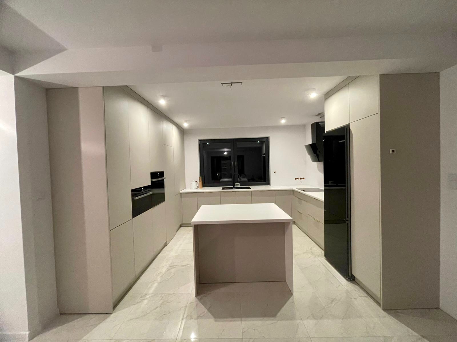 Nowoczesna kuchnia z wyspą i zabudową w jasnych odcieniach szarości, z czarnymi akcentami sprzętu AGD i marmurową podłogą.