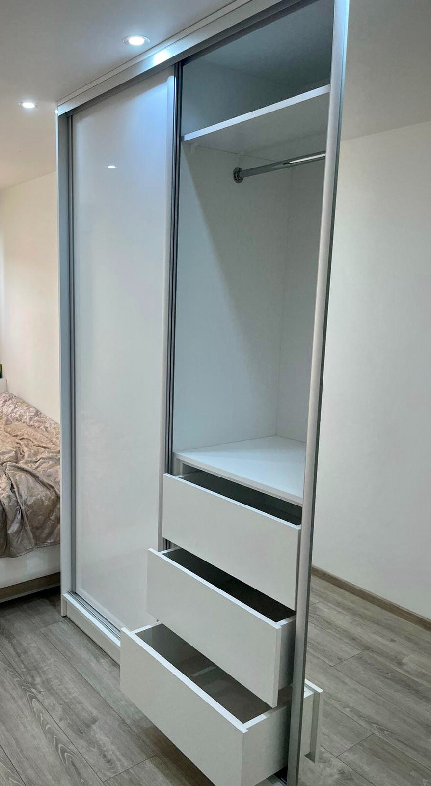Biała szafa wnękowa z przesuwnymi drzwiami i trzema otwartymi szufladami, widoczna półka i drążek na ubrania, podłoga z paneli.