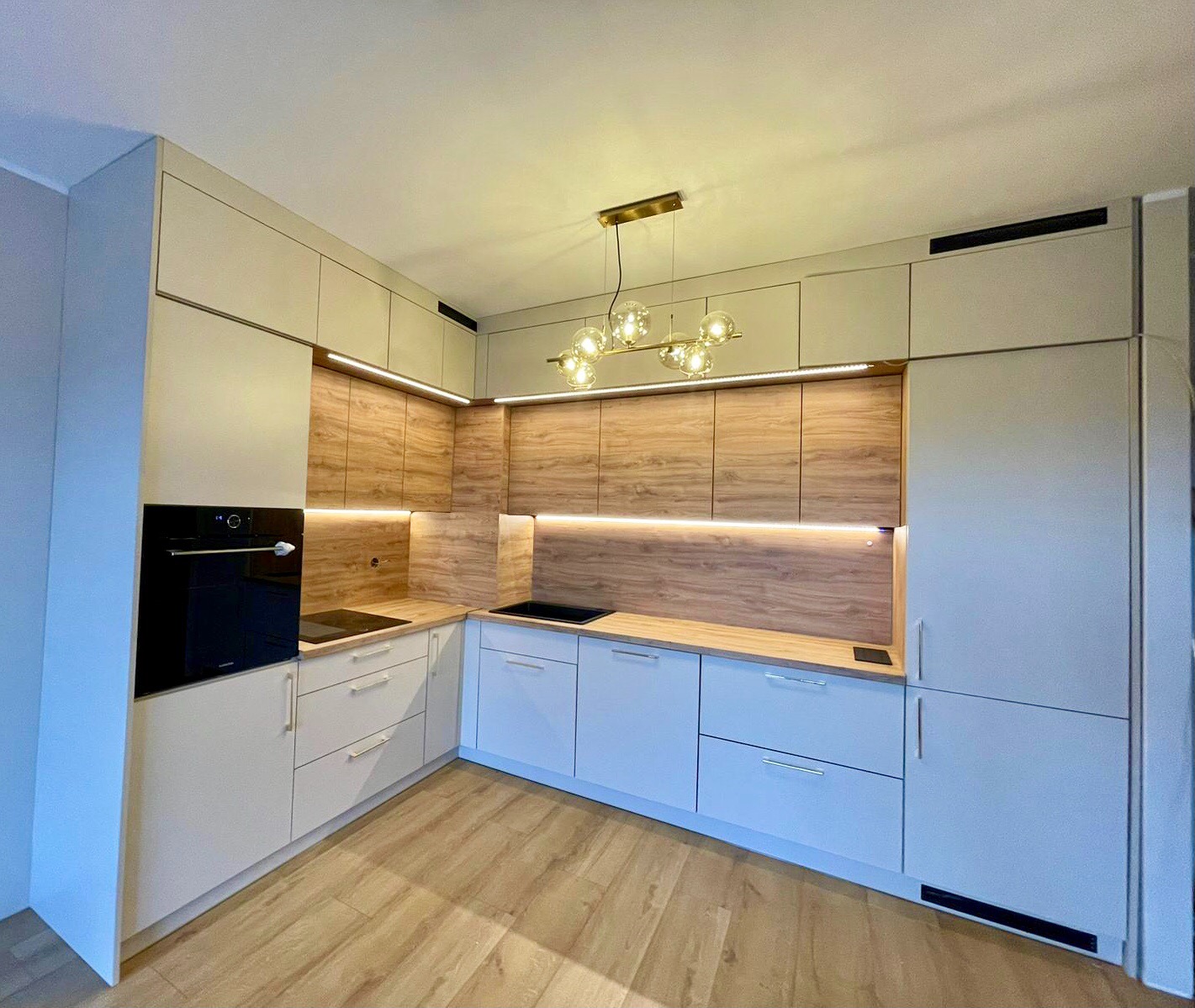 Nowoczesna kuchnia na wymiar z drewnianymi akcentami i oświetleniem LED w Przedborzu. Widoczne białe szafki dolne i górne, drewnopodobne panele ścienne oraz czarny piekarnik.