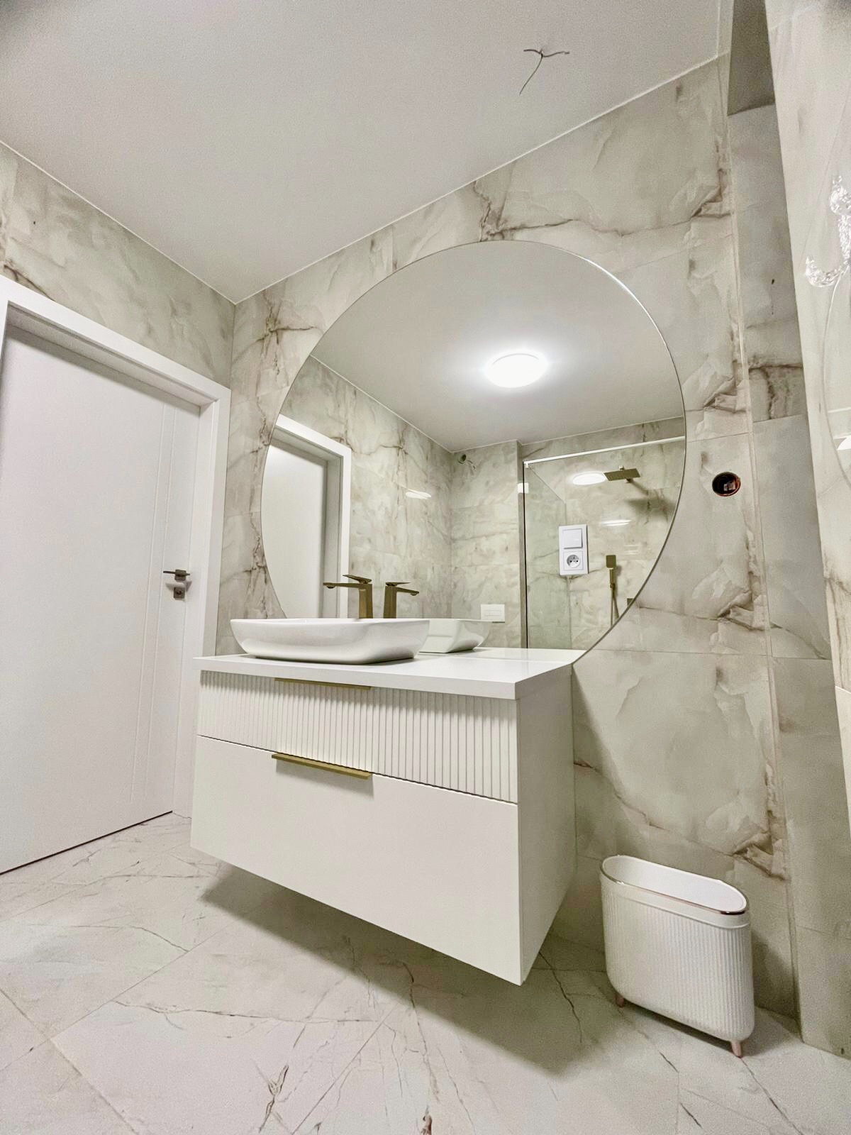 Nowoczesna łazienka w Przedborzu z marmurowymi płytkami, okrągłym lustrem i białą szafką pod umywalkę z frezowanymi frontami oraz złotymi akcentami.