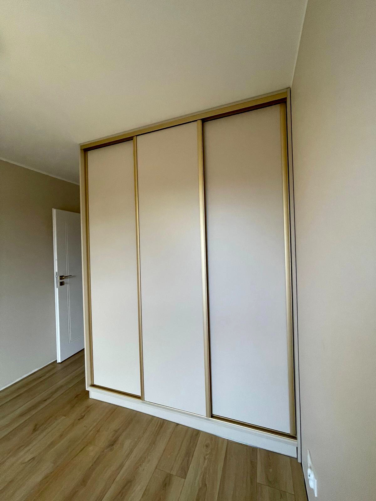 Trzydrzwiowa szafa wnękowa z przesuwnymi drzwiami w kolorze jasnego beżu, z ramami w kolorze złotym, zamontowana w rogu pokoju z drewnianą podłogą i białymi drzwiami w tle.