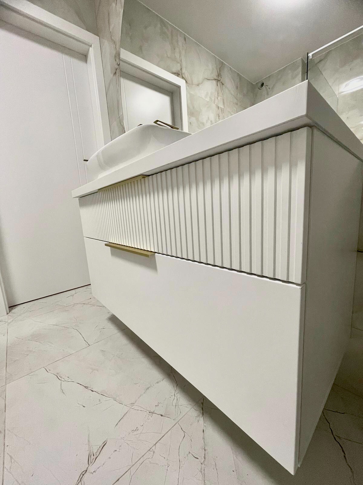 Biała szafka łazienkowa z umywalką nablatową, frezowany front szuflady, złote uchwyty, marmurowe płytki na ścianie i podłodze.