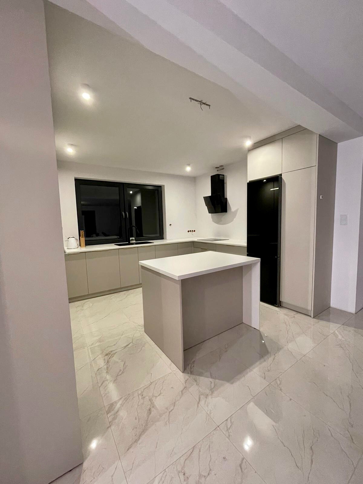 Nowoczesna kuchnia z wyspą, beżowe szafki, czarny okap i lodówka, marmurowe płytki podłogowe.