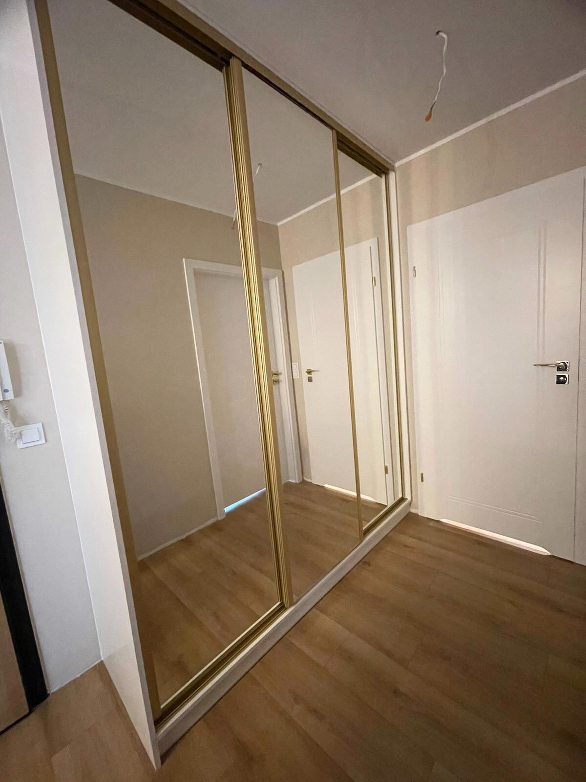 Szafa wnękowa z przesuwnymi lustrzanymi drzwiami w złotych ramach, widoczna w korytarzu z drewnianą podłogą i białymi drzwiami.
