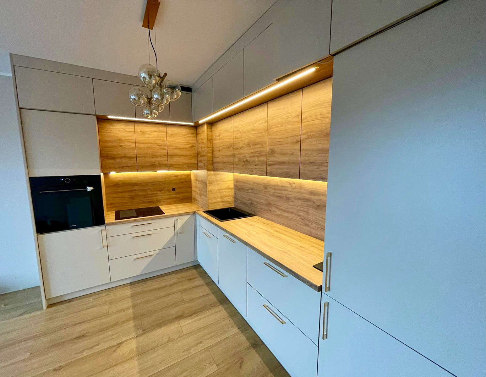 Nowoczesna kuchnia z białymi frontami i drewnopodobnym blatem, oświetlenie LED pod szafkami wiszącymi, szklany żyrandol w stylu retro, płyta indukcyjna i czarny zlew.