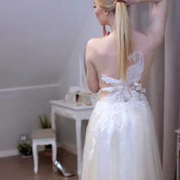 Kobieta w sukni ślubnej z odkrytymi plecami, ozdobionymi aplikacją w kształcie pawia, poprawia włosy na tle toaletki z lustrem.