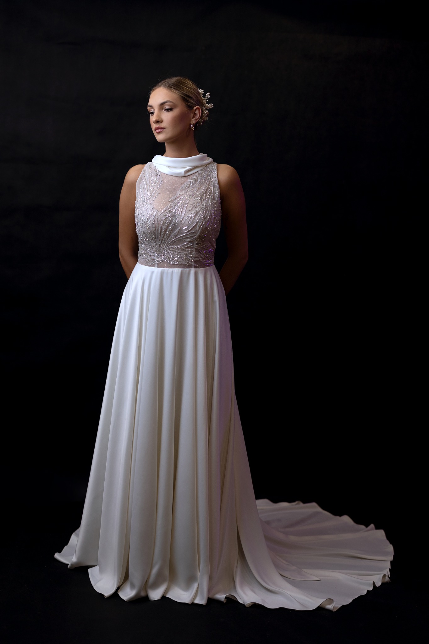 Elegancka kobieta w długiej, białej sukni z bogato zdobionym, prześwitującym gorsetem i delikatnym upięciem włosów z biżuteryjnymi akcentami na ciemnym tle.